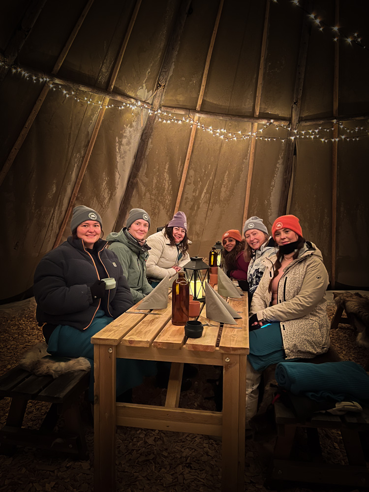 Women in a ski lodge socialising at a table with drinks and warm lights at night, winter holiday.