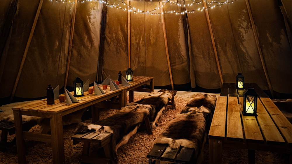 Warmly lit camping tent with wooden tables and benches, lanterns and fur rugs inside Sweden