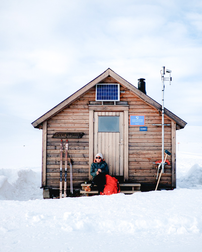 Woman by a wooden cross-country ski cabin in snowy landscape, with skis and winter gear outside