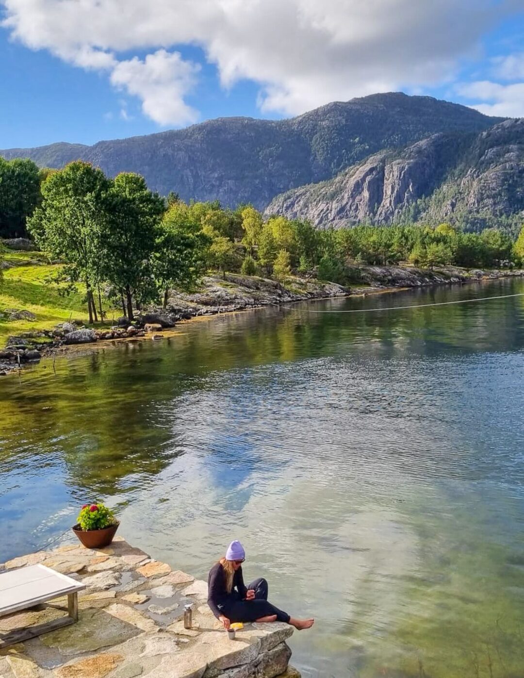 Woman sitting by a calm riverbank in a mountain valley landscape, with greenery and blue sky overhead