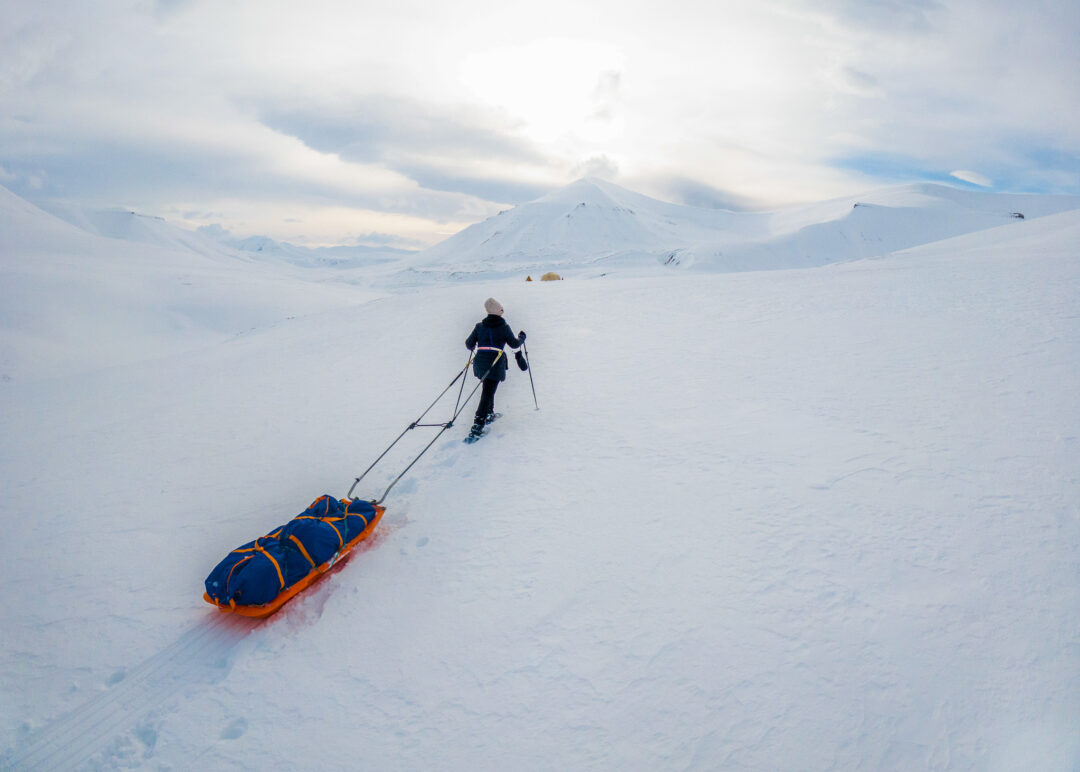 Woman snowshoeing across snowy Arctic landscape, pulling a sled trail in the mountains