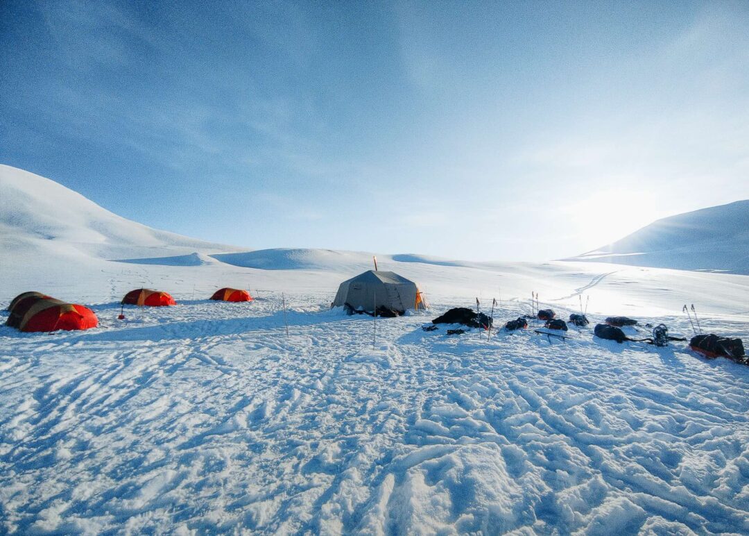 Wild camping in the Arctic: tents pitched on a snowy landscape with distant mountains and trekkers