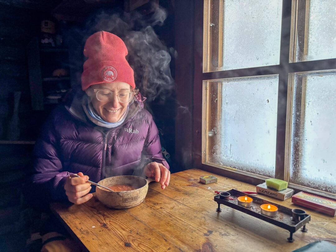 Woman in winter clothing cooking soup in a cabin by a stove near a window