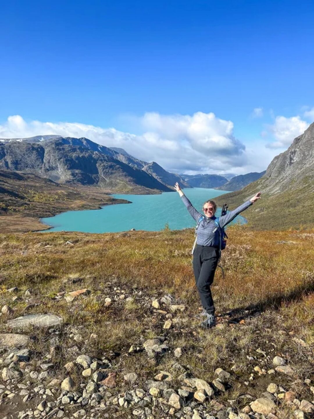 Women hiking with arms out on a trail above a turquoise fjord in Norway, surrounded by mountains.