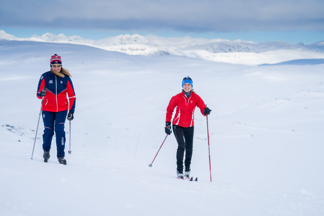 Women cross-country skiing on snowy mountains in Norway, wearing red ski jackets and skis on a trail.