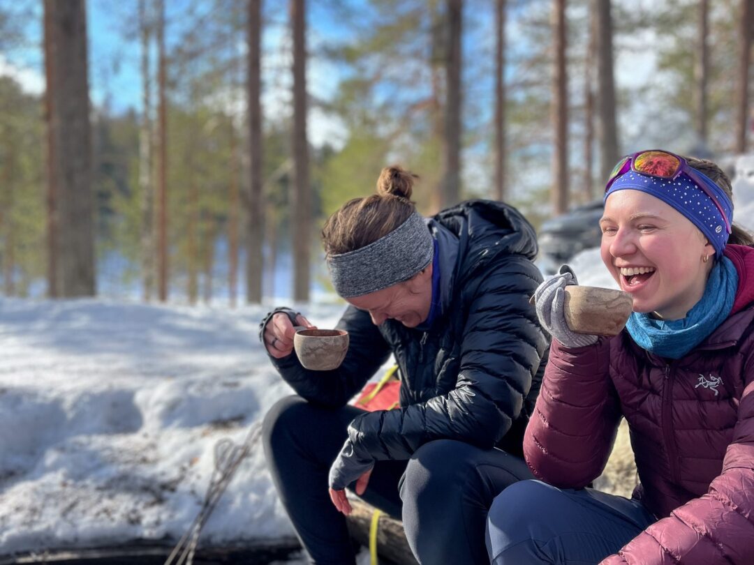Women cross-country skiing, laughing and sharing hot drinks in a snowy forest trail.
