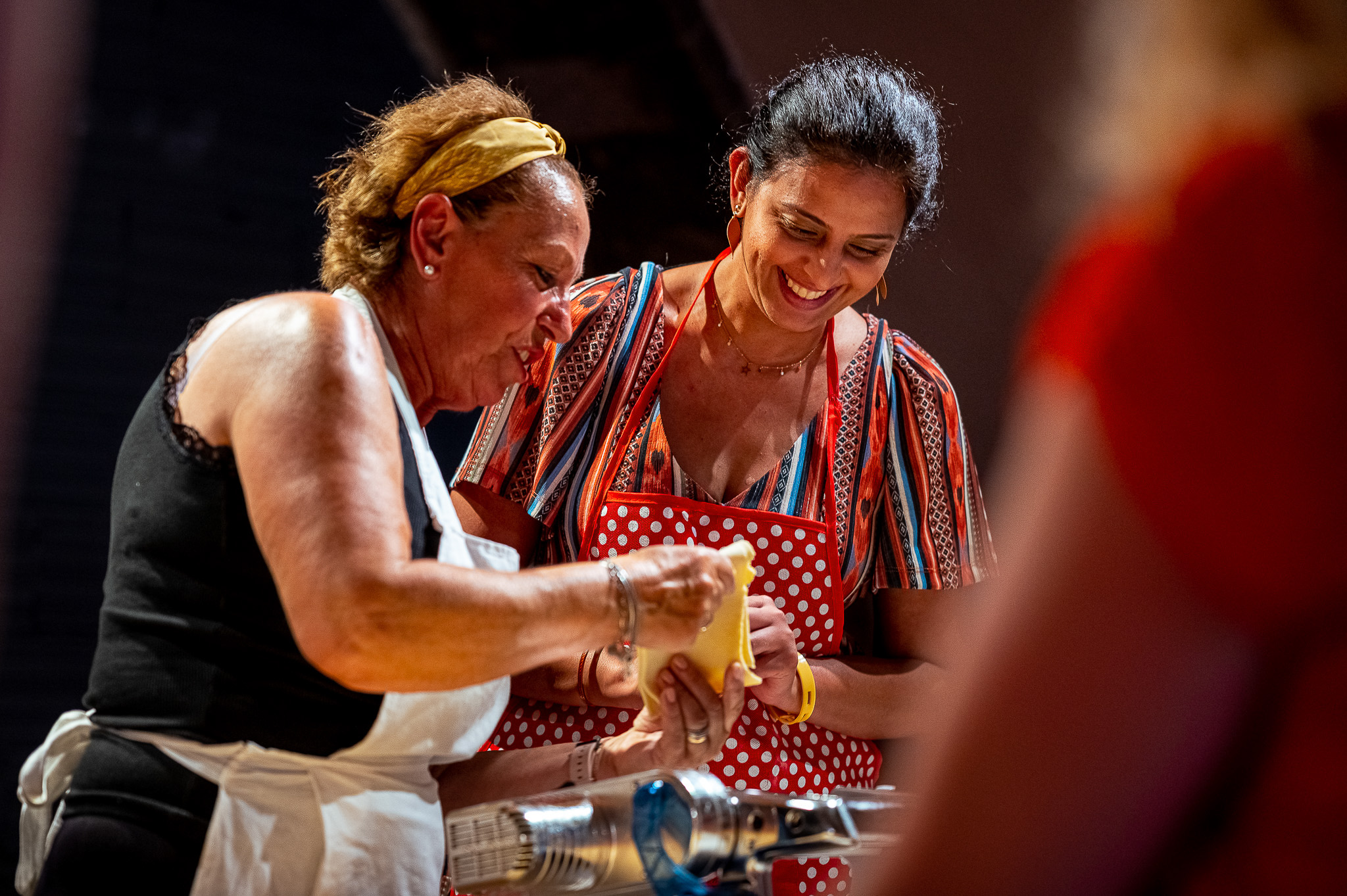 Two women cooking together, smiling, wearing colourful aprons indoors