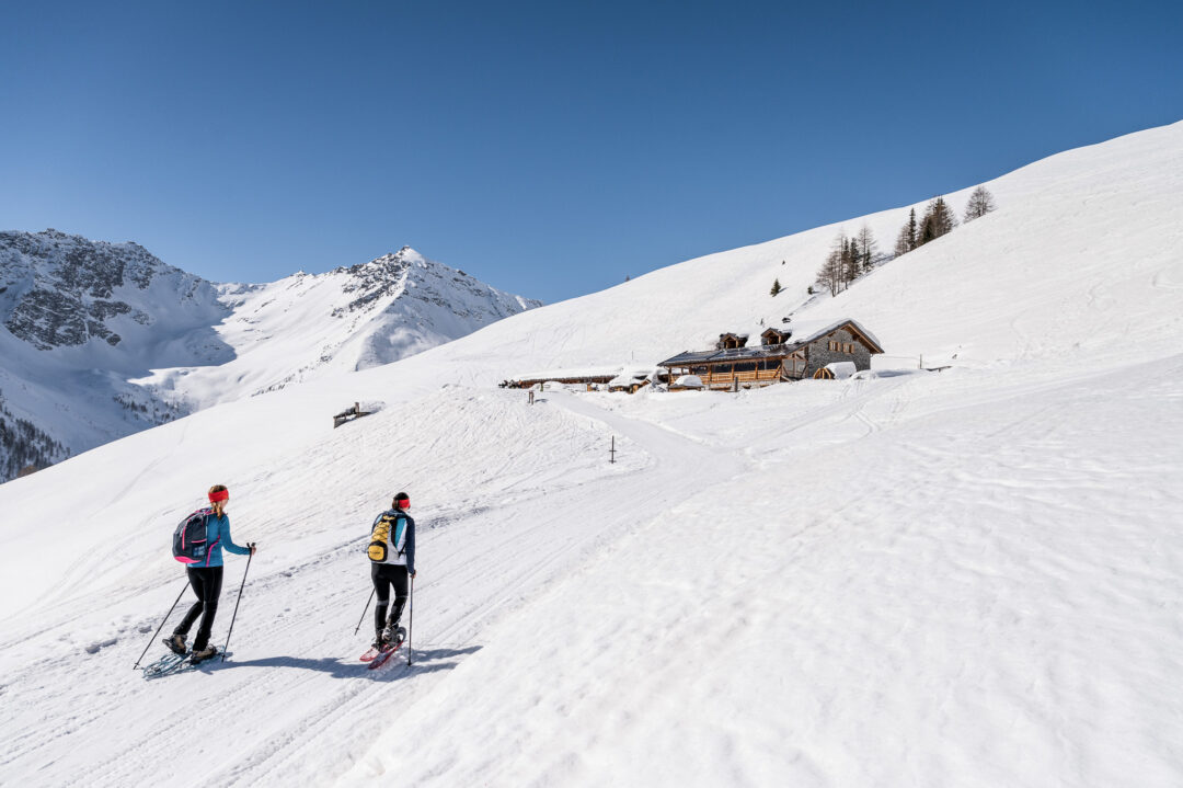 Women snowshoeing across snowy alpine slopes past mountain huts under a clear blue sky
