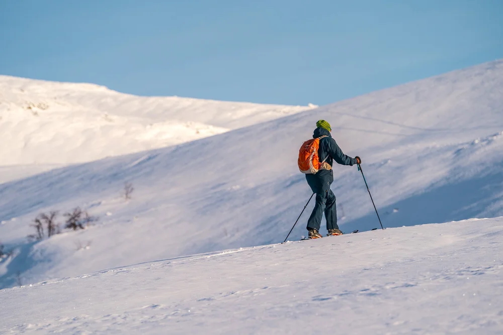 Woman ski touring uphill on snowy mountains in Norway, with poles under a clear blue sky