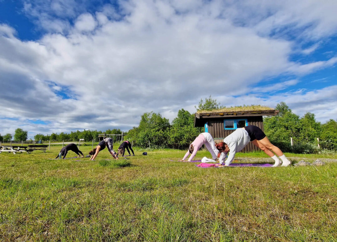 Women doing outdoor yoga on mats in a grassy field near a wooden barn with horses in the distance
