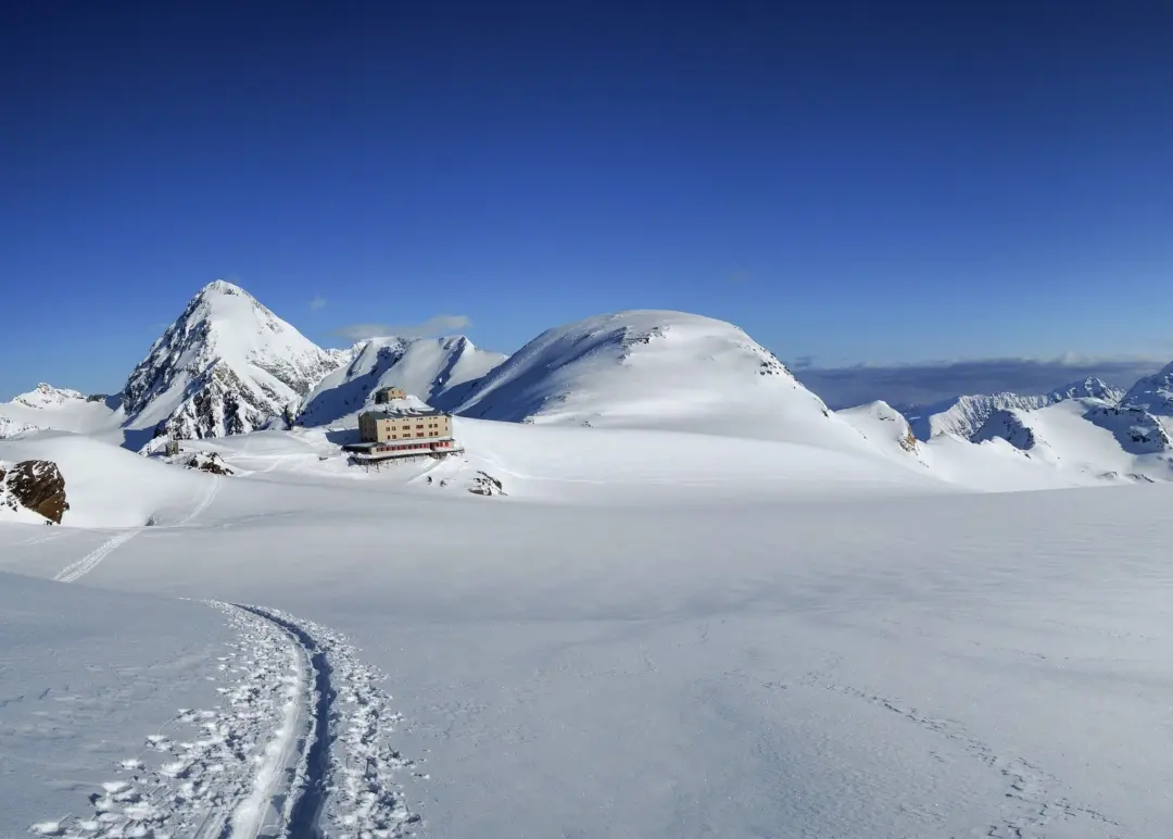 Snow-covered mountain landscape with winding trail tracks and a small chalet under a clear blue sky