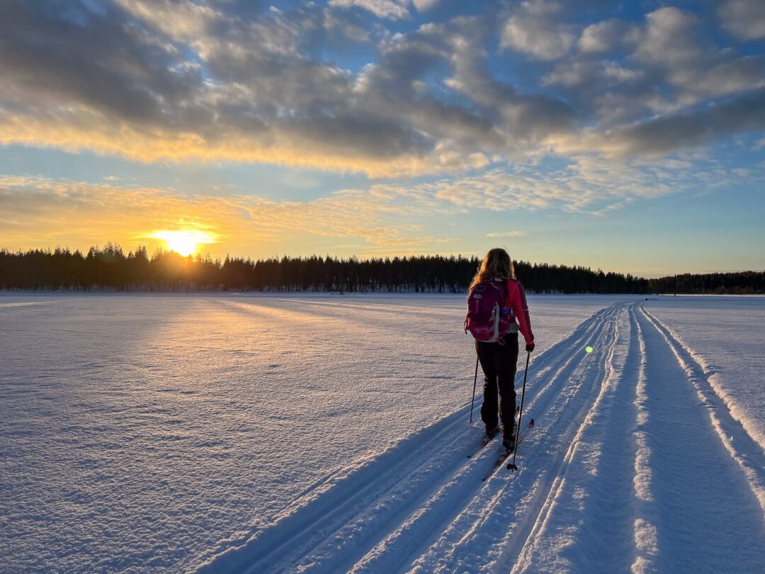 Woman cross-country skiing on a snowy trail across Finland at sunrise, with a winter forest backdrop.