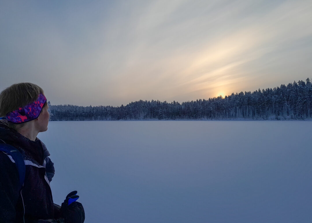 Woman in winter jacket watching snowy Arctic landscape at dawn over a frozen lake and forest