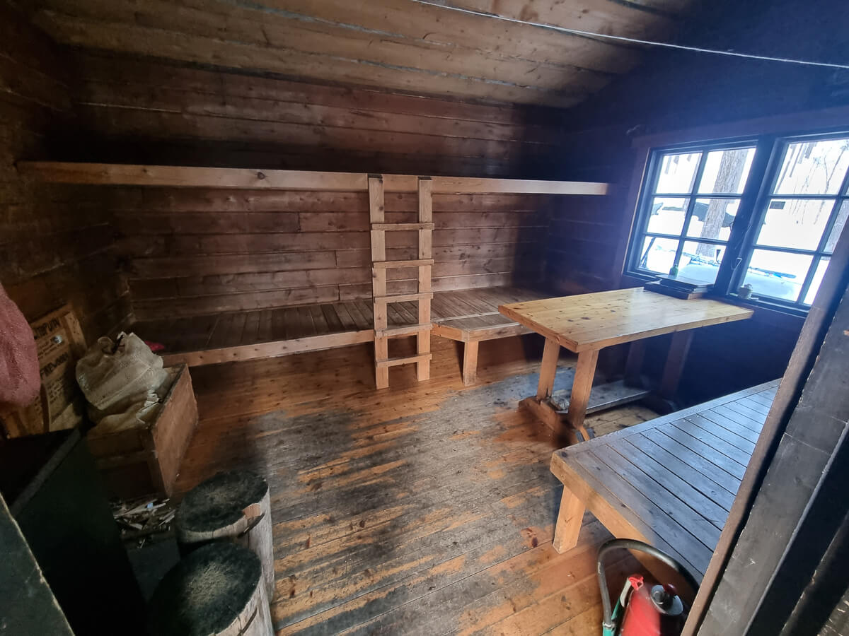 Wooden backcountry ski hut interior with benches, table and window, showing skis and boots.