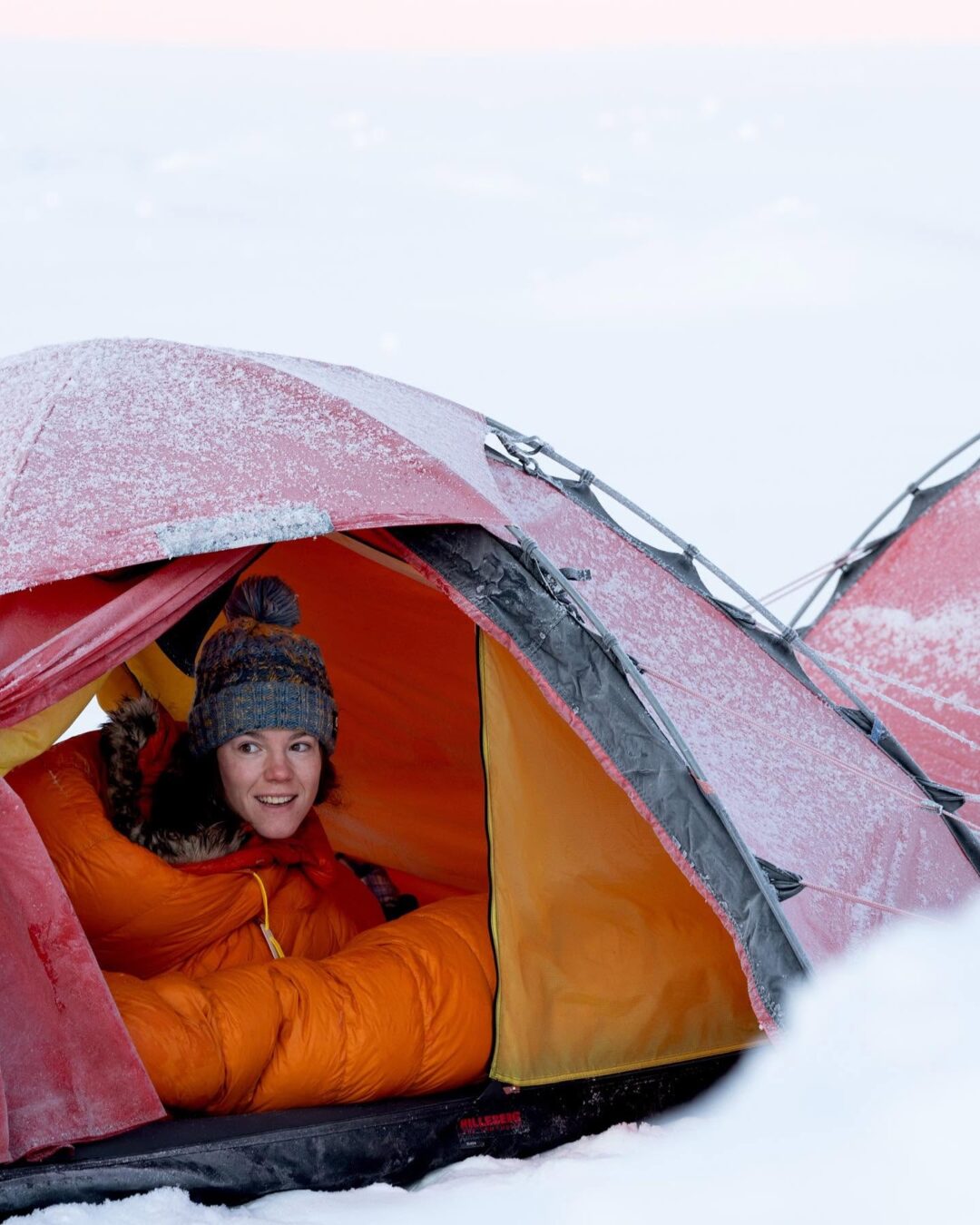 Woman inside a winter camping tent in snowy conditions, wearing a warm hat and orange jacket