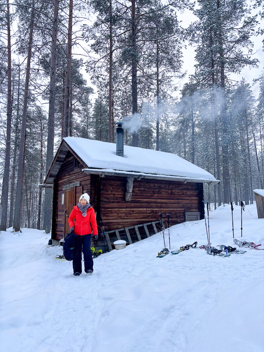 Woman in snowy forest by a wooden cabin, with cross-country skis lined up outside