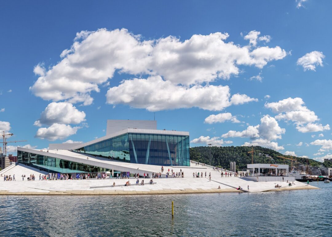 Oslo Opera House on the waterfront, with visitors walking the sloped roof under a blue sky and clouds