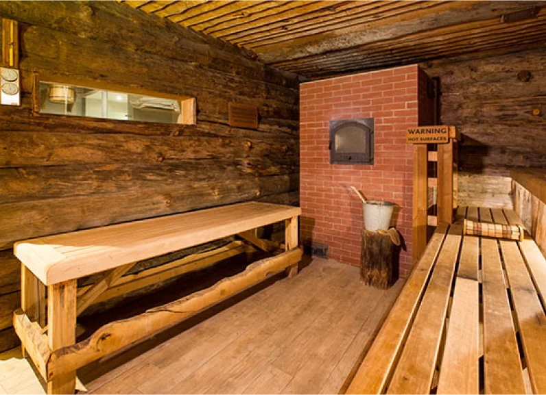 Wooden sauna interior with brick stove, benches and “WARNING HOT SURFACES” sign.