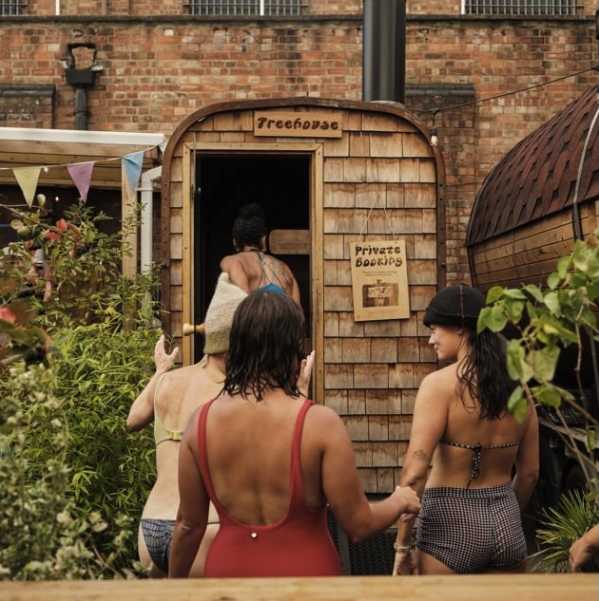 Women entering an outdoor wooden sauna in a courtyard, with signage reading “Private Bookings”.