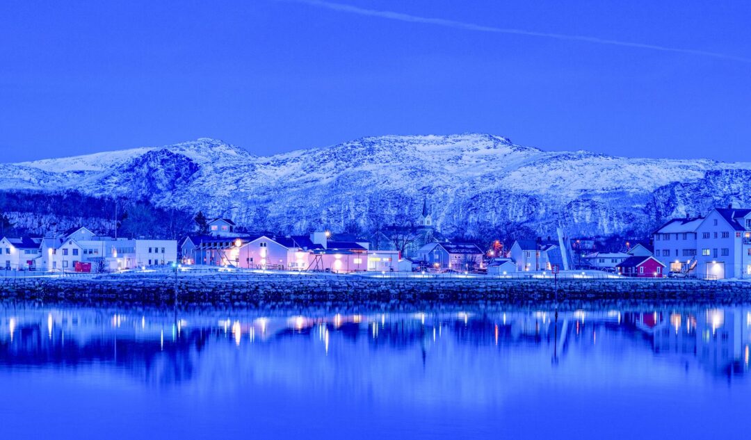 Snowy Arctic winter village by a calm fjord at night with bright lights reflected in the water