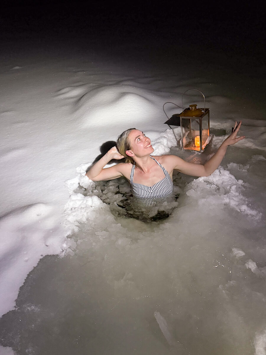 Woman sits in icy water at night, arms out beside a lantern during winter camping.