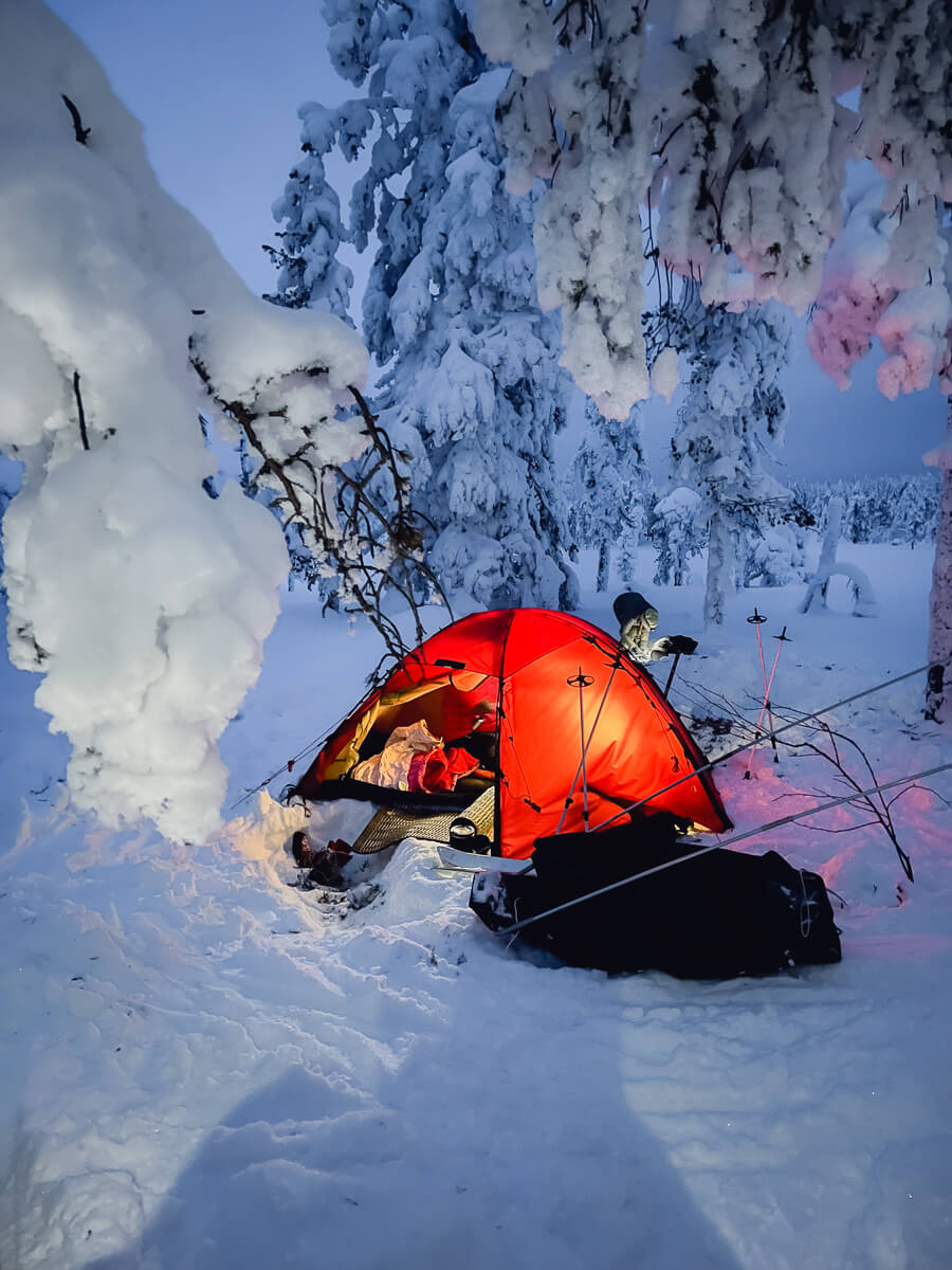 Red and orange backcountry winter camping tent in a snowy forested mountain landscape