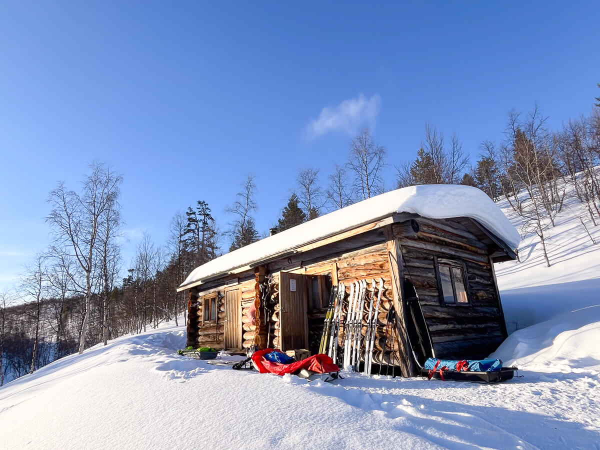 Wooden backcountry ski hut in snowy forest with skis and expedition gear outside under blue sky