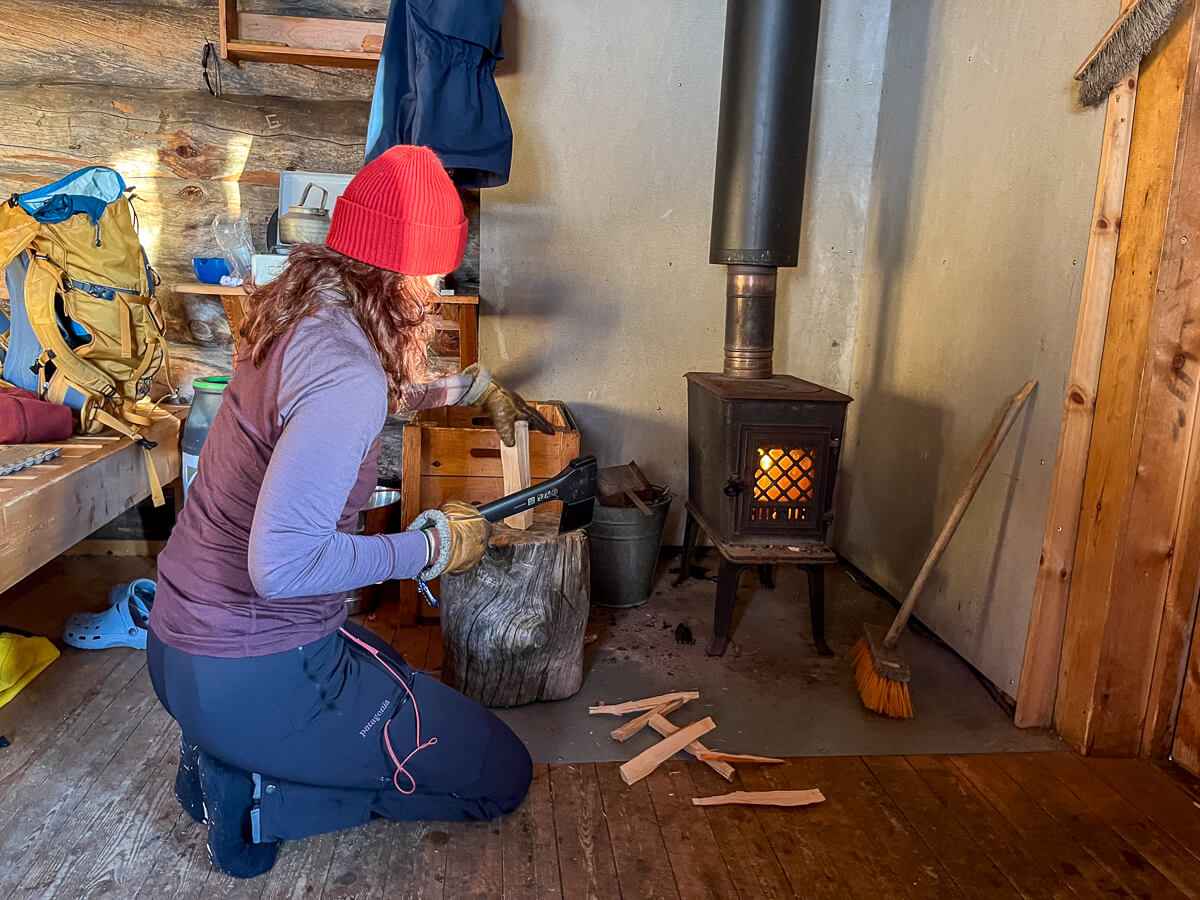Woman chopping kindling by a wood stove inside a backcountry ski hut.