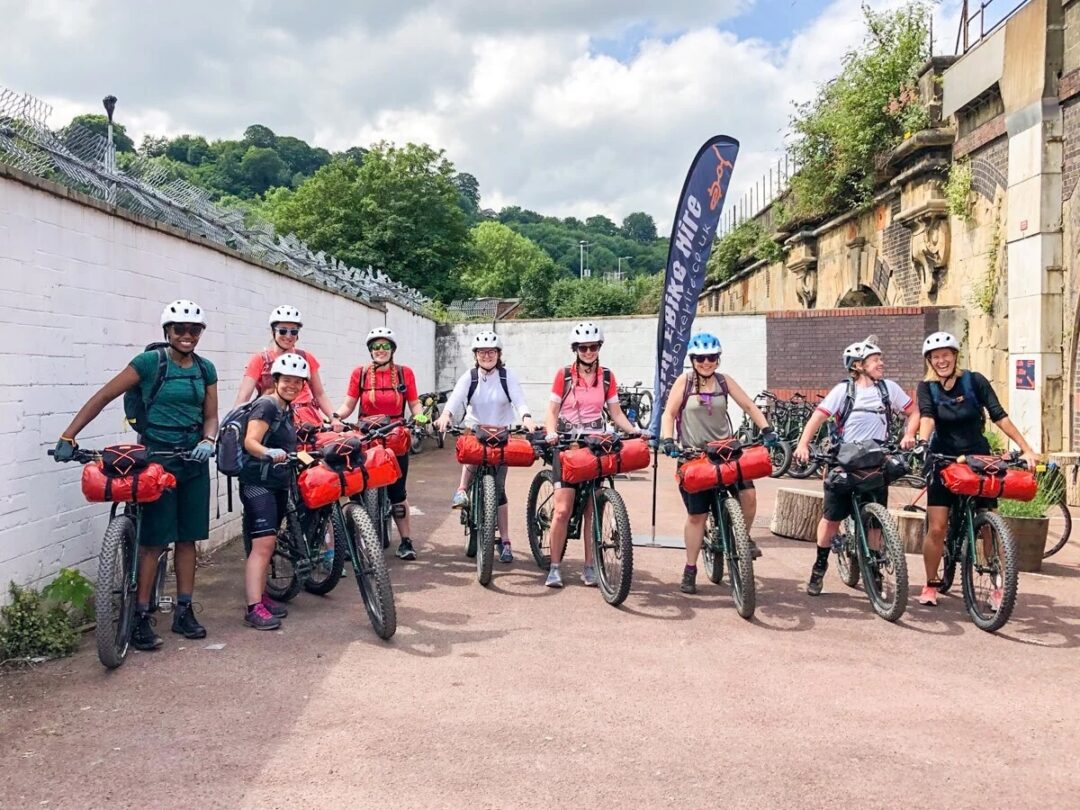 Women bikepacking with bicycles and red panniers outside stone buildings in Bath, wearing helmets.
