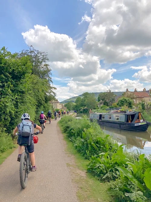 Women cycling along a canal towpath past narrowboats under cloudy skies