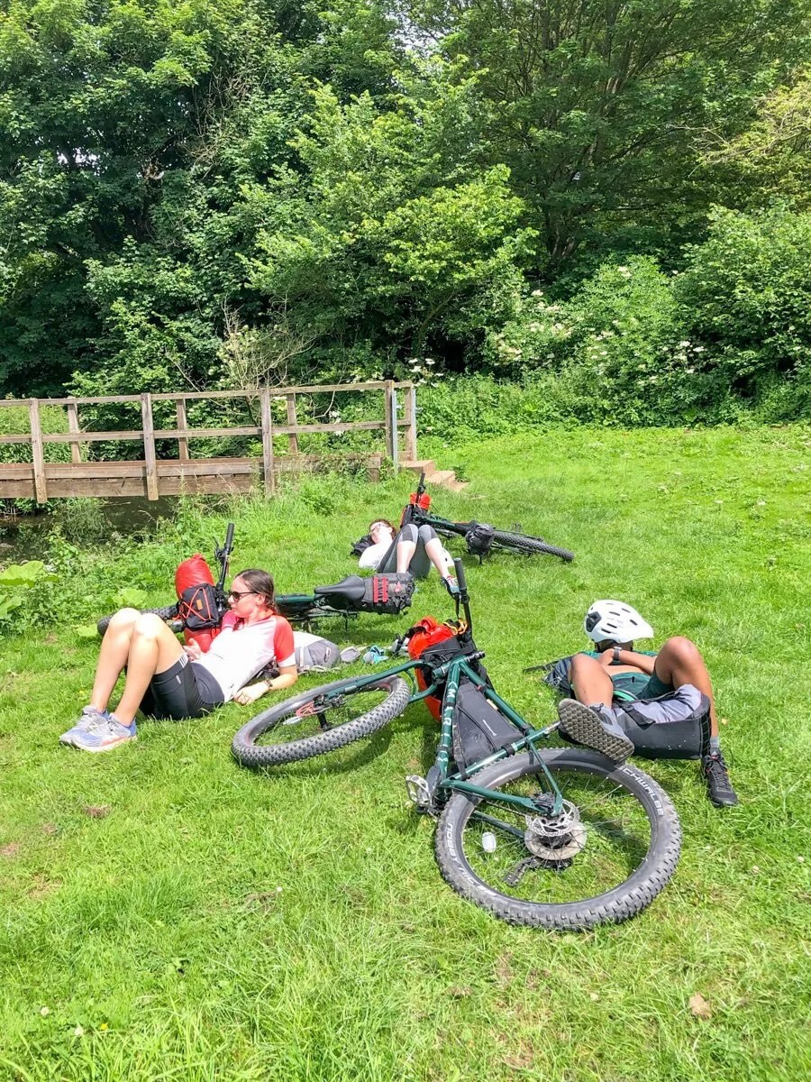 Women bikepacking on a grassy riverside stop, with bikes parked beside them in lush green countryside.