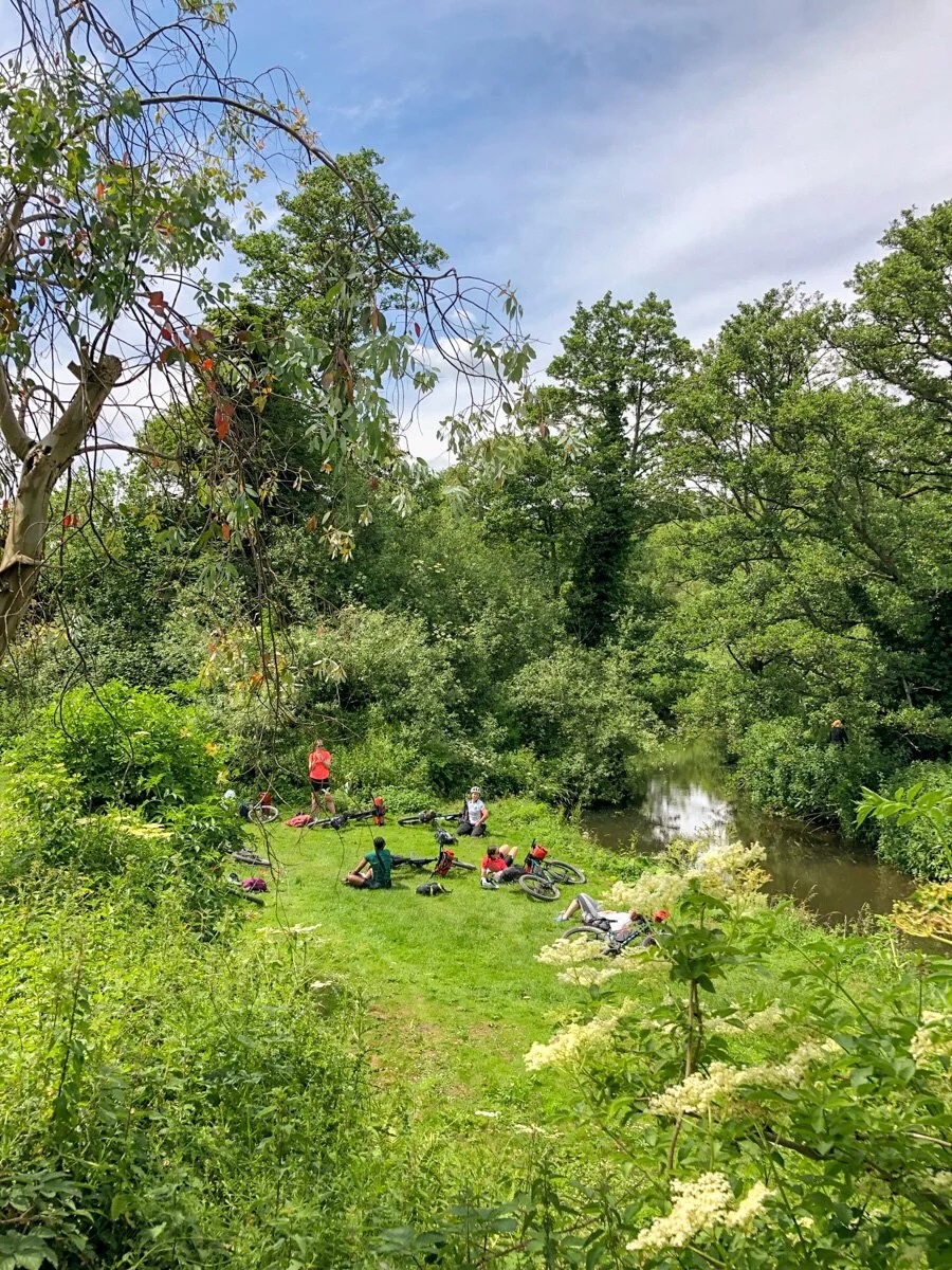 Cyclists picnic on grass beside bikes by a river in a park, Bath.