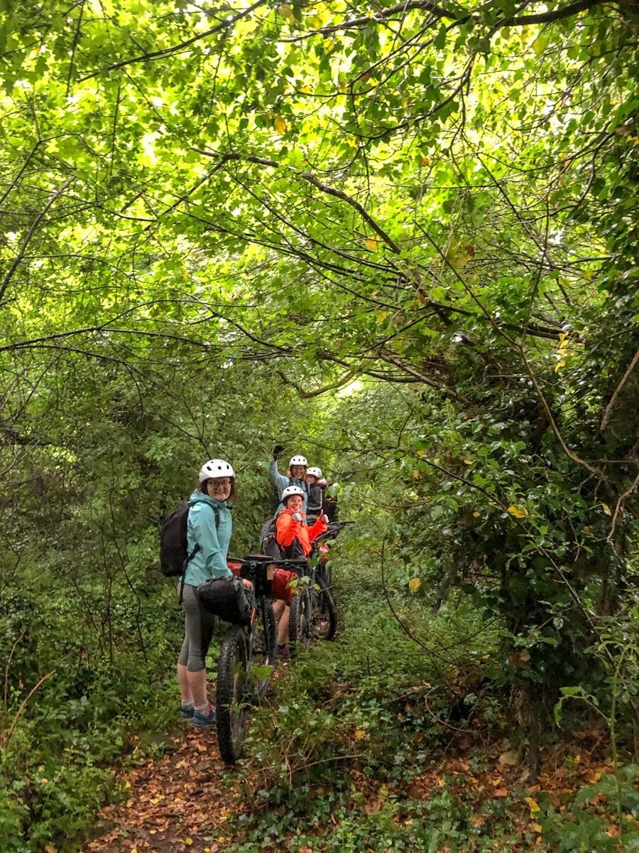 Women bikepacking riders in helmets on a wooded trail, surrounded by green spring branches.