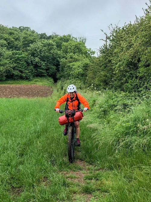 Women cycle on a grassy trail through green countryside near Bath, wearing helmets and bikepacking gear