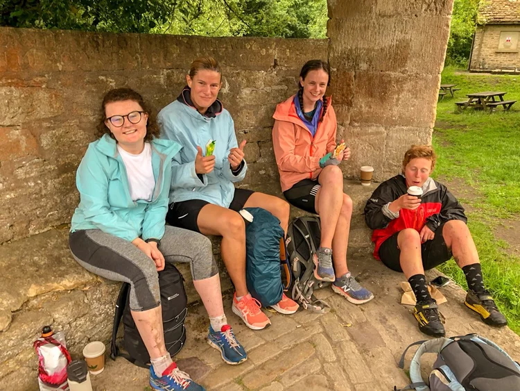 Women on a bikepacking break, sitting outside by a stone wall with backpacks and cycling gear.