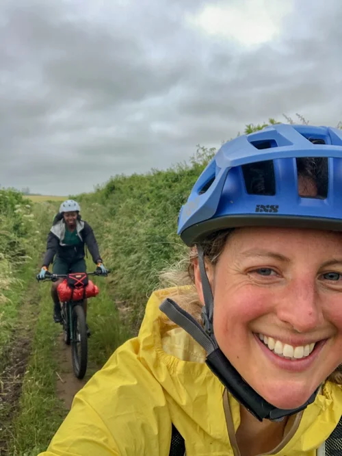 Smiling woman cyclist in a helmet on a gravel path, with another woman biking in the background