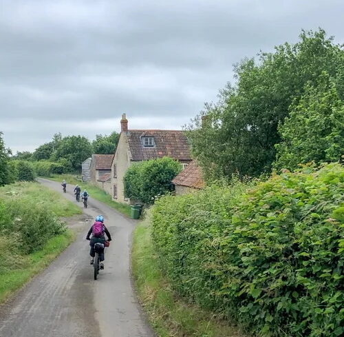 Women bikepacking on a quiet country road past cottages, surrounded by trees and greenery