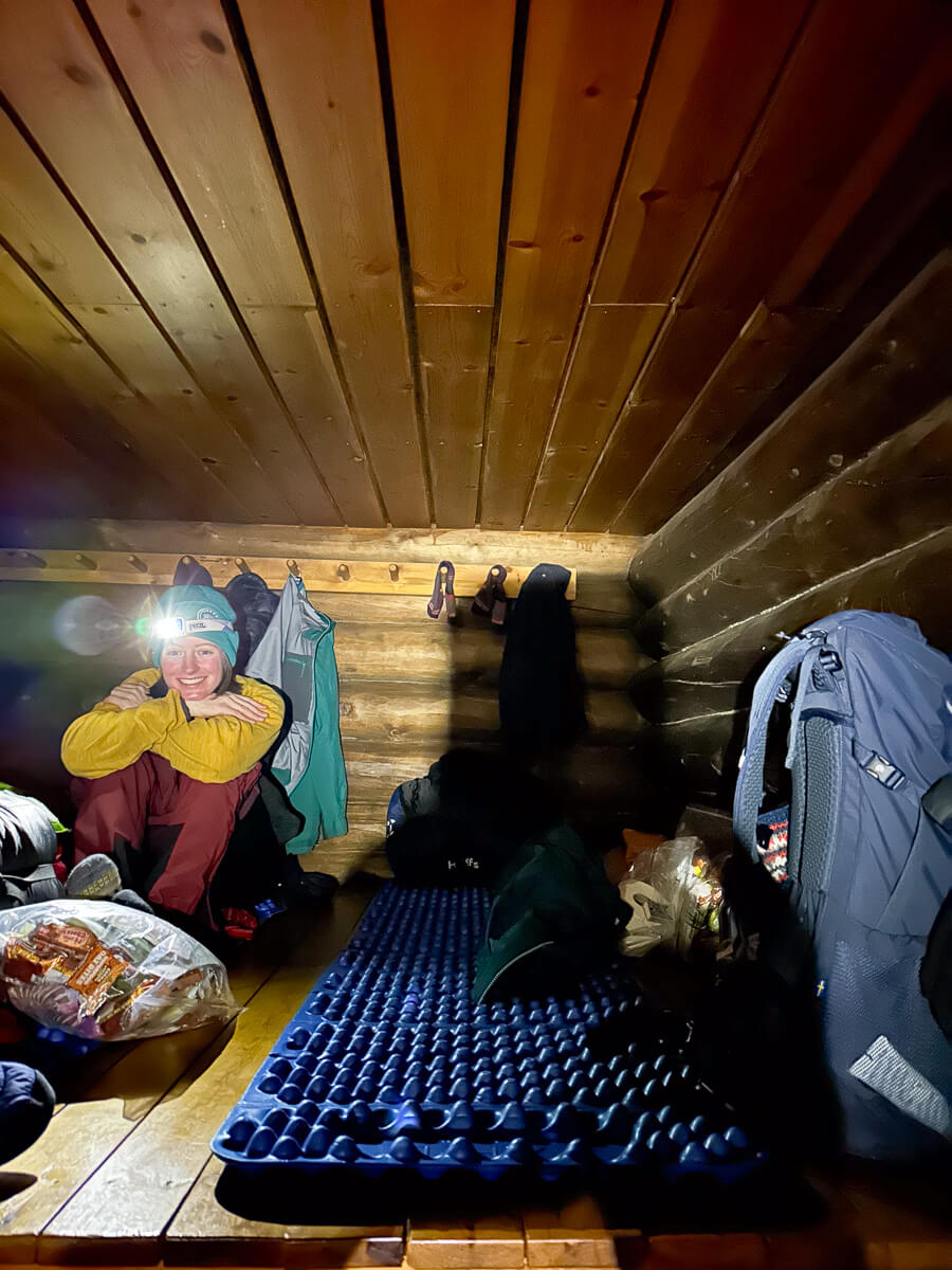 Woman in winter kit inside a backcountry ski hut in Lapland, with camping gear and bunks