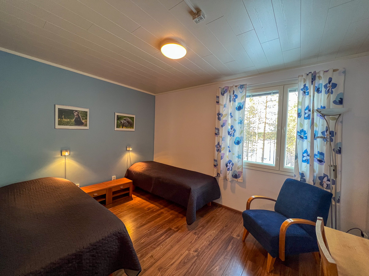 Well-lit bedroom with three beds, chairs and a large window with blue floral curtains.