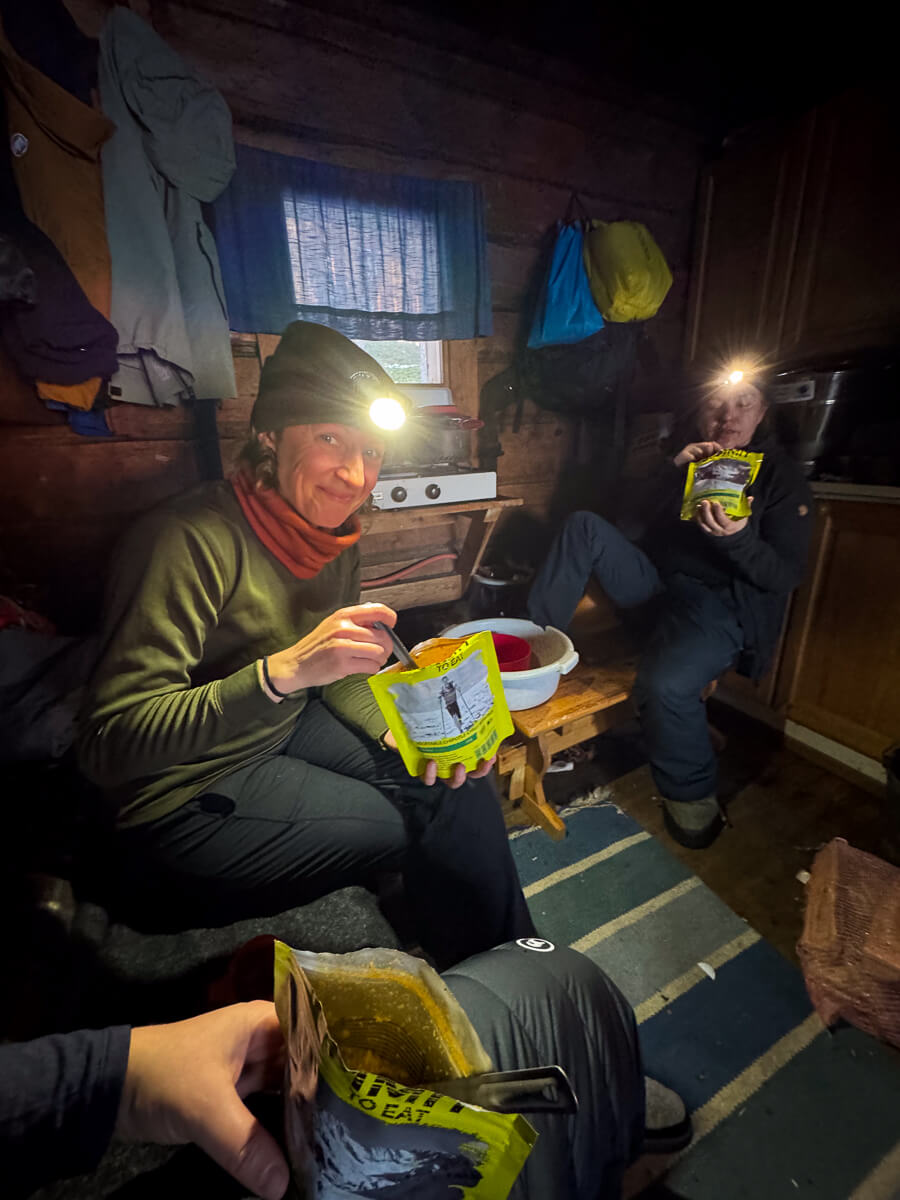 Women inside a backcountry ski hut at night, eating snacks and holding food packets.