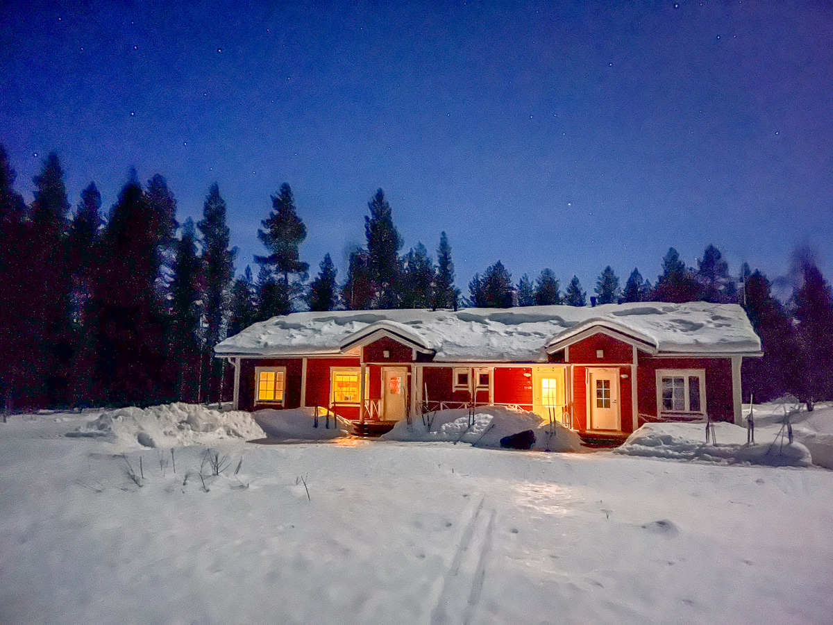 Snow-covered cabin in a Finnish winter landscape at dusk, with warm lights glowing in the snow.