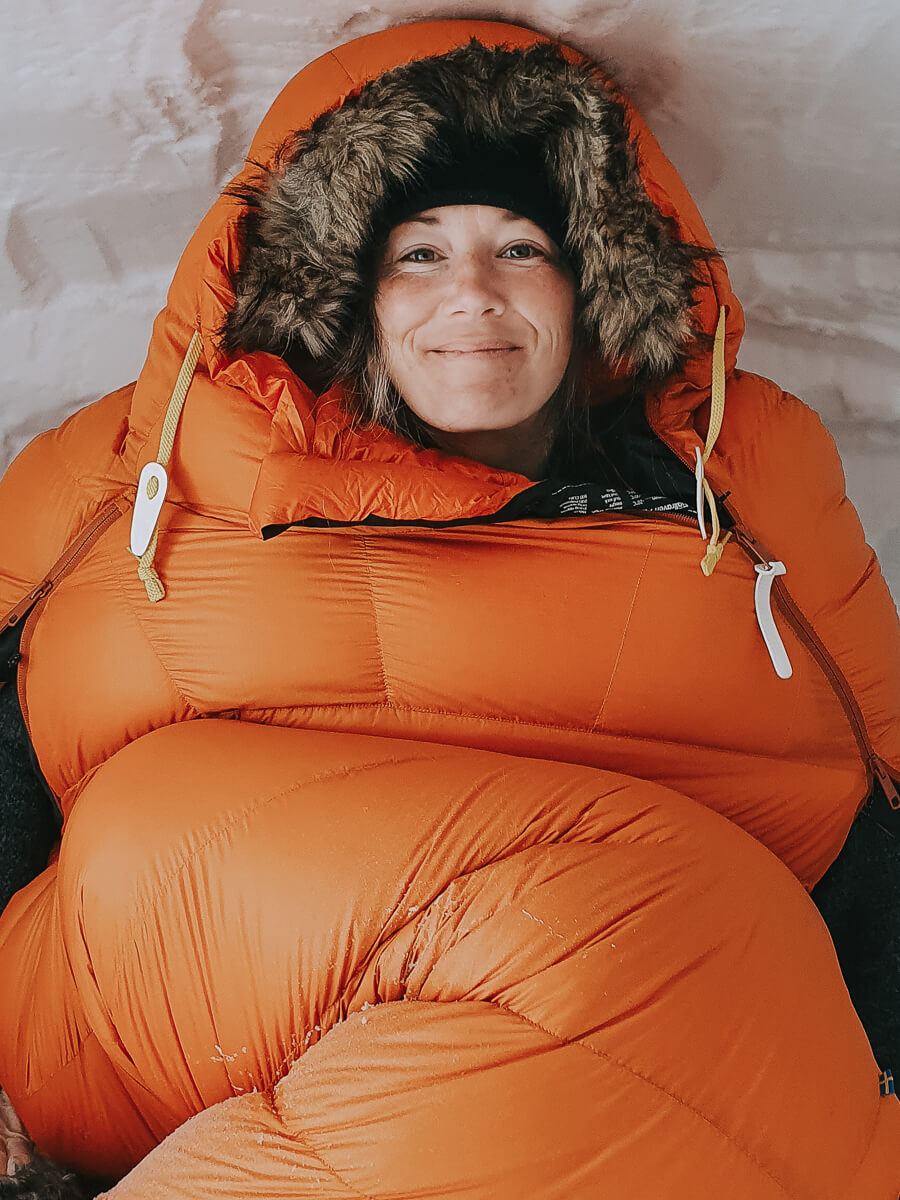 Woman smiling in an orange sleeping bag hood during winter camping in snowy conditions