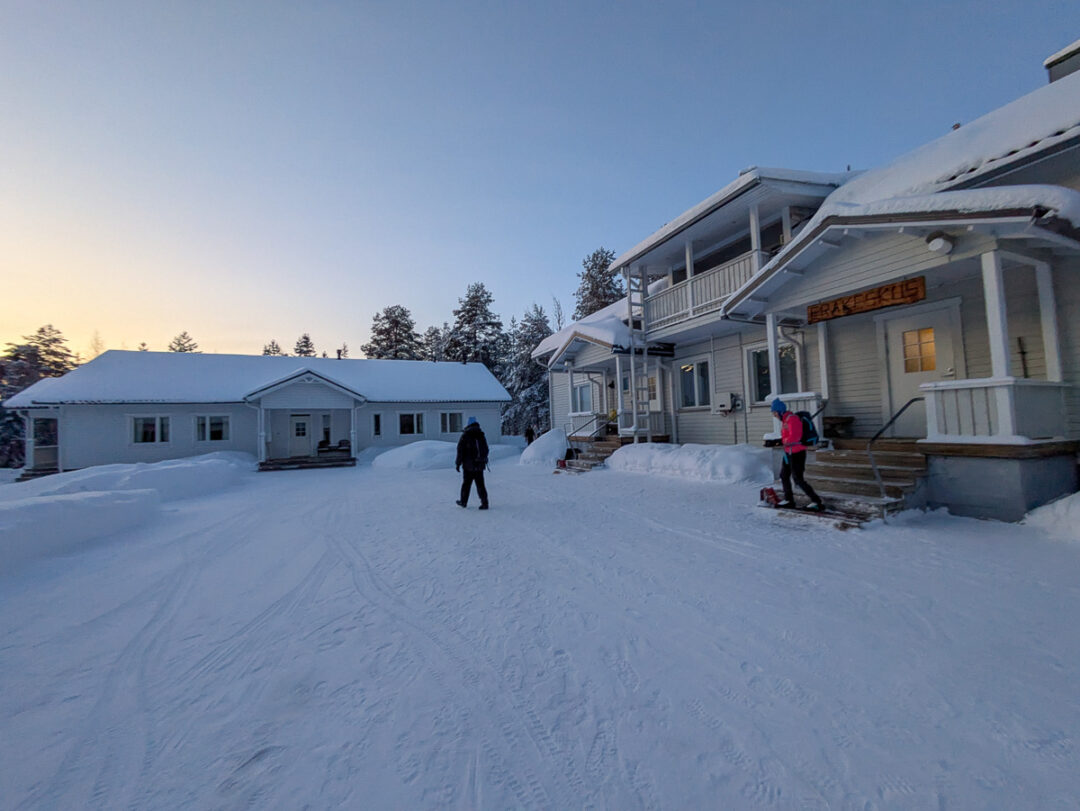 Cross-country skiers on snowy tracks beside winter lodge buildings at dusk
