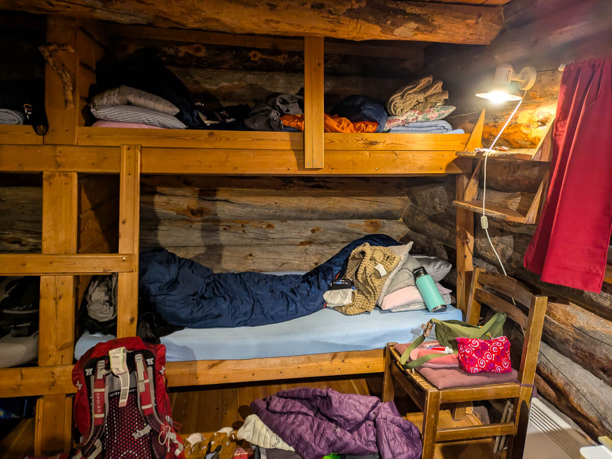 Cozy wooden hut interior with bunks, sleeping bags and camping gear beside a lamp
