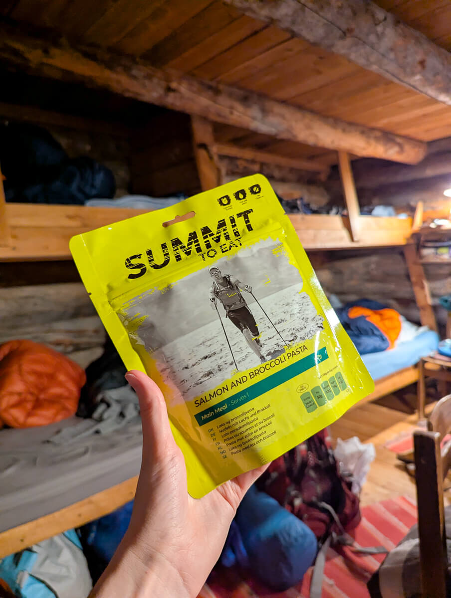 Hand holds yellow “Summit to Eat” pouch for salmon and broccoli pasta inside a wooden ski hut