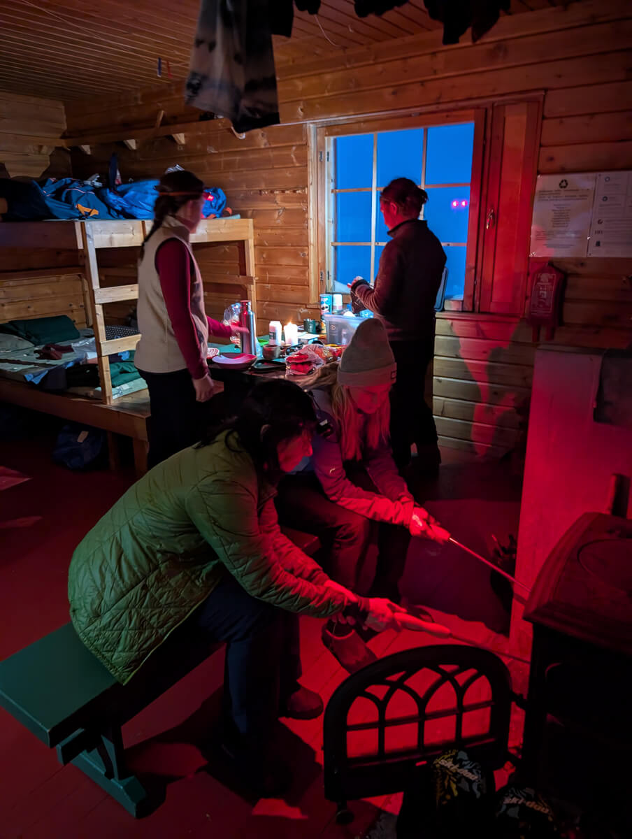 Women warming up in a wooden backcountry ski hut by a stove, night interior with winter light through window