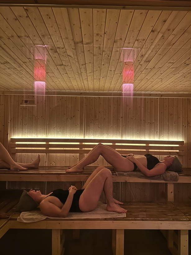Women relaxing in a wooden UK sauna, with pink heat lights above the bench seating