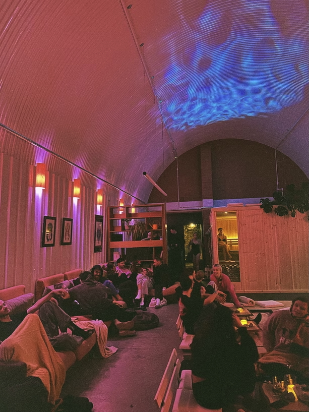Guests relaxing in a heated sauna lounge with pink and blue lighting, seated on benches with drinks