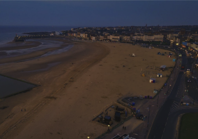 Aerial view of a UK beach at dusk with a seafront promenade and sand, with tents and huts along the shore