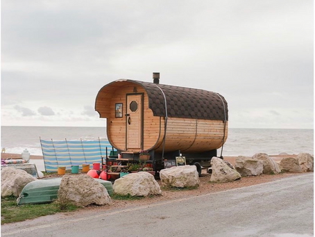 Barrel sauna by the sea with a wooden outdoor cabin on a rocky shoreline, with camping gear nearby.